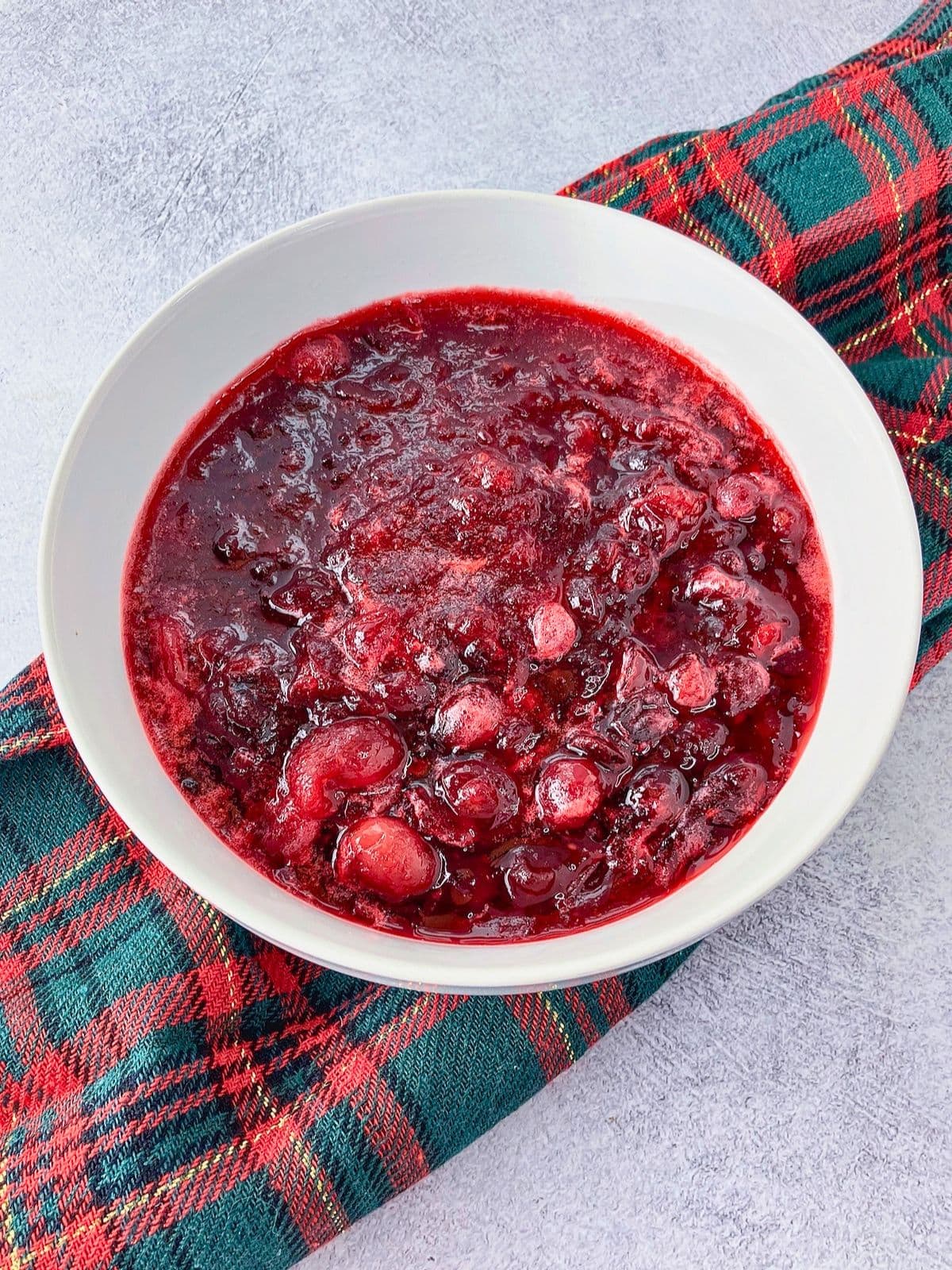 Homemade cranberry sauce with whole and burst cranberries is served in a white bowl on a red and green plaid cloth. The sauce has a thick glossy texture and deep ruby red color.