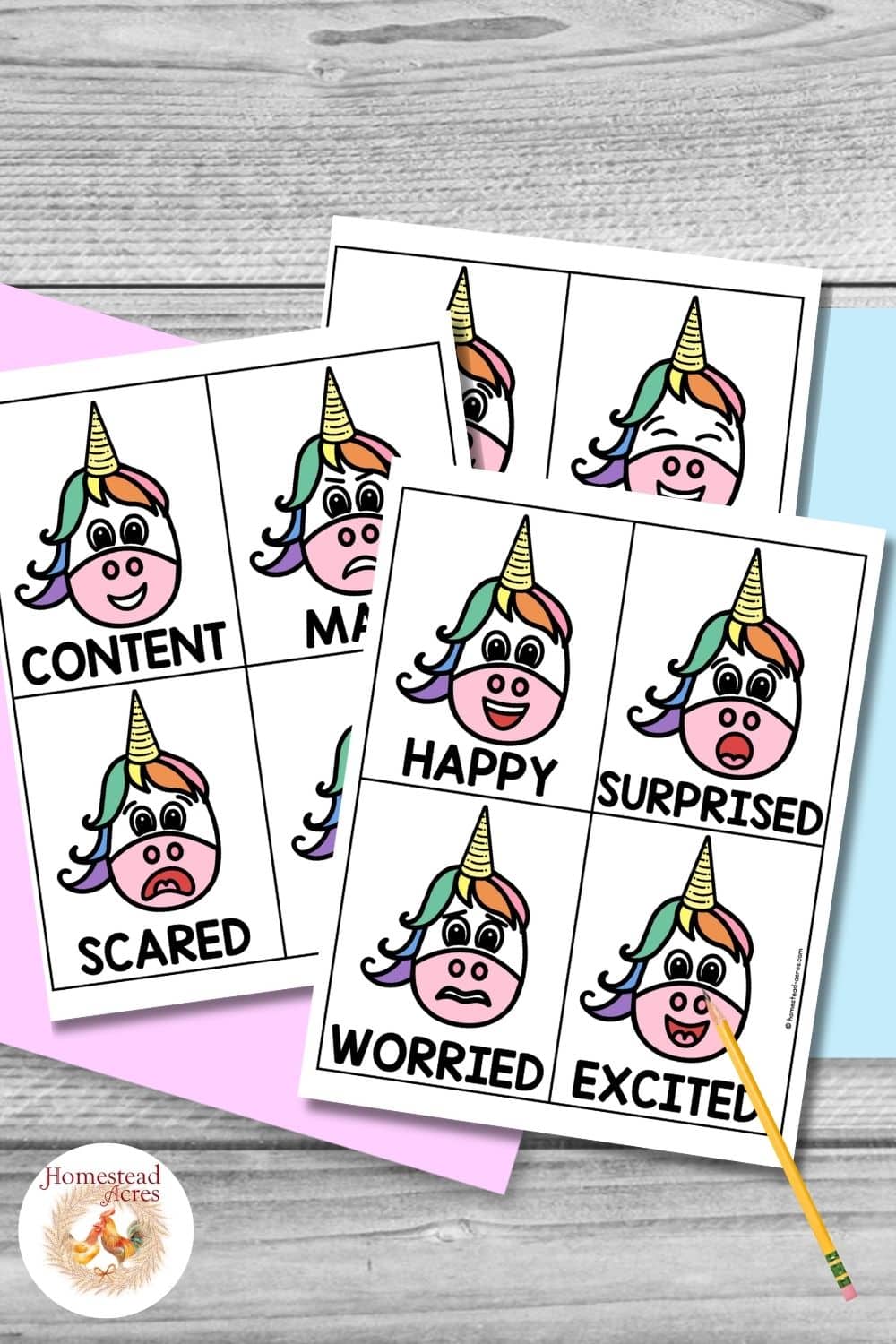 Color unicorn emotion cards arranged on pastel paper with a pencil on a gray wood background. Visible feeling labels include "CONTENT". "MAD". "SCARED". "HAPPY". "SURPRISED". "WORRIED". "EXCITED".