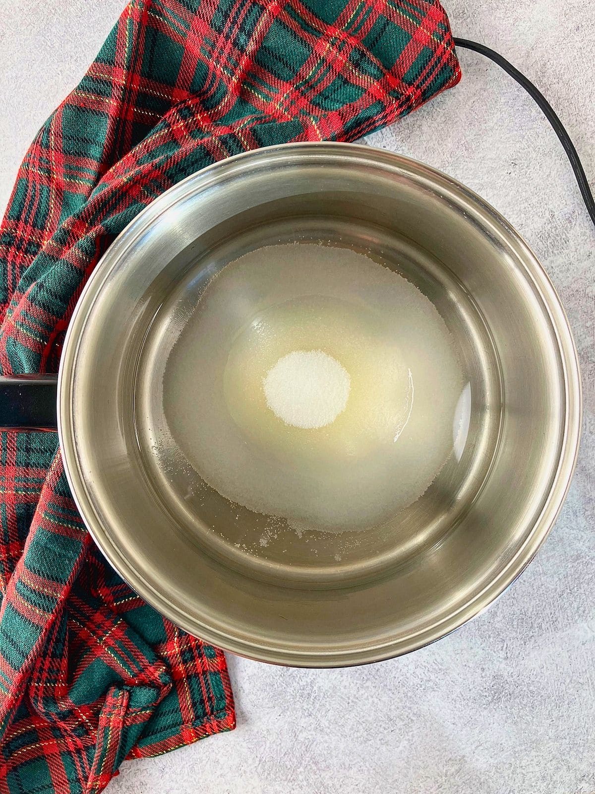 Water and granulated sugar sit in a stainless steel saucepan, ready to be heated. A red and green plaid cloth rests beside the pan.